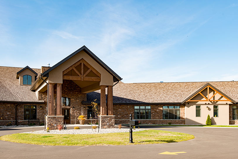 Ballard Glenn Assisted Living facility exterior in Appleton, Wisconsin — modern timber-frame entrance, rustic stone facade, landscaped circle drive under blue sky.